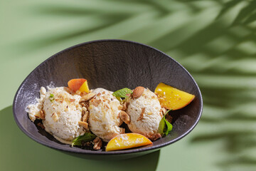 three scoops of ice cream with nuts and peach slices on a dark plate on a green background with a shadow from a palm tree, top view space for text