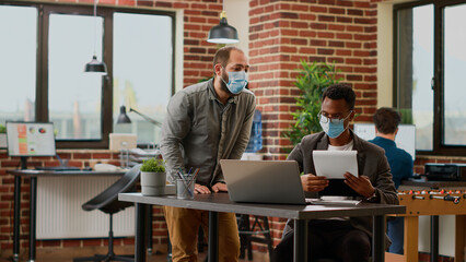 Office workers planning business presentation in startup boardroom, talking about research information and charts analysis. People with face masks working with documents and laptop.
