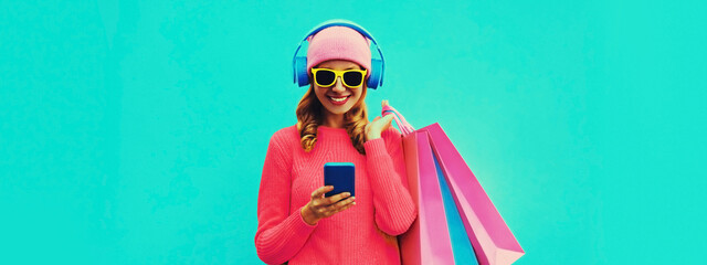 Farbenfrohes Porträt einer stilvoll lächelnden jungen Frau, die Musik über Kopfhörer hört, mit Einkaufstaschen, die mit Strickpullover und rosa Hut auf blauem Hintergrund posieren