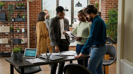 Diverse group of coworkers planning research analysis in business office, working on startup partnership to create project report with charts data and statistics. Using documents and digital tablet.