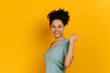 Excited happy african american curly haired young woman in a t-shirt, points finger to the side behind the back, at empty copy space, stands on isolated orange background, looks at camera, smiling