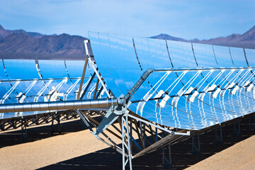 Curved mirrors of a solar heat collectors in the desert with mountains and blue sky.
