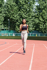 Focused sportive black woman enjoys training on sunny summer day preparing for upcoming marathon. Young African American athlete runs on red track of stadium against city park with lush green trees