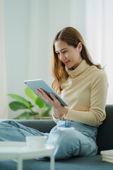 Asian woman holding a tablet on the sofa at home doing financial transactions and online shopping.