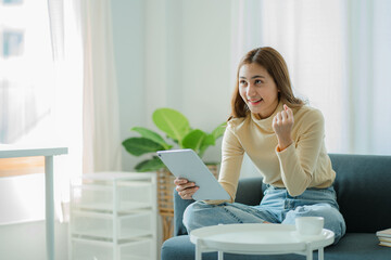 Asian woman holding a tablet on the sofa at home doing financial transactions and online shopping.