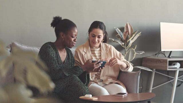 Medium slowmo of two multiethnic female colleagues looking at smartphone and talking, working together in marketing agency