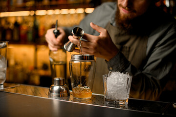Close-up view on glassy shaker into which pouring drink from jigger