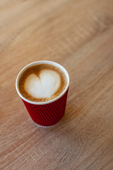 Cup of cappuccino with latte art on wooden table. Beautiful foam, red paper cup. Coffee to go on wooden background with heart shaped latte art on top. Morning breakfast. Take away.