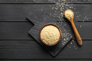 Board with bowl and spoon of sesame seeds on dark wooden background
