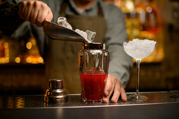 close-up of shaker with drink into which bartender pours ice cubes using scoop