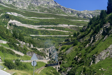 Look up to serpentine mountain road of Swiss Grimsel Pass with smoke of Furka steam train on a sunny summer day. Photo taken July 3rd, 2022, Oberwald, Switzerland.