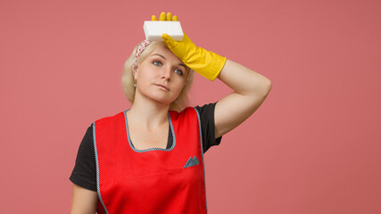 tired maid with a sponge in her hand wipes sweat from her forehead on a pink background
