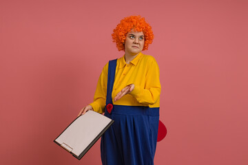 funny clown in a wig and a yellow-blue suit is perplexed and holds a clipboard in his hand on a colored background