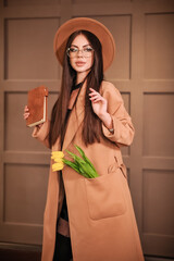 Portrait of a beautiful stylish brunette in round glasses with make-up in a brown hat and raincoat stands indoors yellow tulips flowers stick out of her pocket and in her hands a notebook