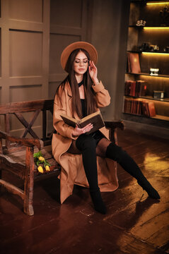 A beautiful stylish brunette in round glasses with makeup in a brown hat and a raincoat sits on a bench indoors and reads a book and yellow flowers tulips lie nearby