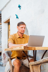 Bearded caucasian man using laptop outside, sitting at wooden table in hotel. Male hands typing on laptop keyboard. Concept of young people working on mobile devices on vacation.