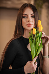 Beautiful stylish brunette woman with makeup holding yellow flowers in her hands stands indoors