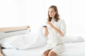 Woman caring of her beautiful skin on the face applying moisturizer on her face. Smiling girl holding little jar of skin cream and applying lotion after shower