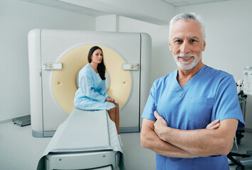 Portrait of experienced doctor standing in medical radiology room with CT scanner, female patient...