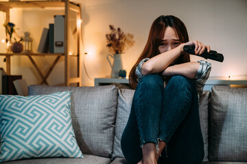 asian young woman sitting huddled on the sofa is burying her face in fear while watching a horror film on tv alone in the living room at home during nighttime.