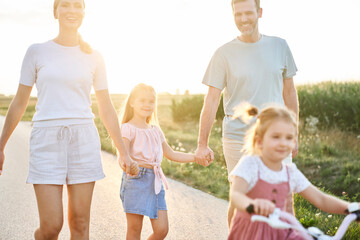 Family of four caucasian people spending time on walking and riding a bike on village road