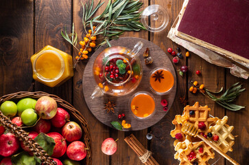 Autumn tea with berries and honey. Sea buckthorn and raspberries in a glass teapot, cups with poured tea. Apples and waffles with honey.