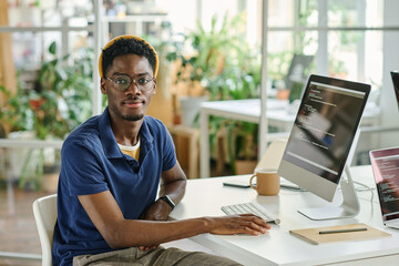 Portrait of African young developer in eyeglasses looking at camera while sitting at his workplace with computer monitor
