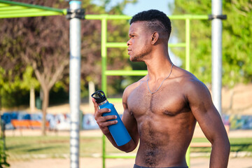 Portrait of a young fit shirtless black man holding water bottle looking at a side in a park...