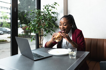 Young successful freelancer African American woman food blogger or vlogger in the cafeteria eating and testing fat tasty American hamburger or burger and give it a score for blog or live web cam video