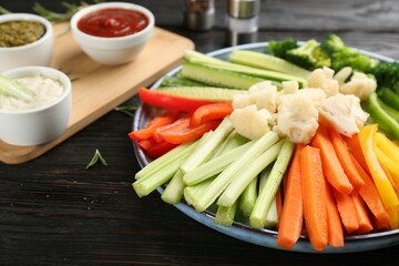 Plate with celery sticks, other vegetables and different dip sauces on dark wooden table, closeup