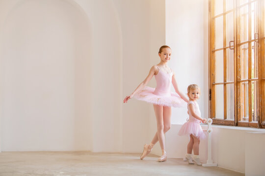 The Older Sister, A Ballerina In A Pink Tutu And Pointe Shoes, Shows The Baby How To Practice .