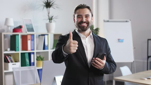 Caucasian man in formal suit looking at smartphone screen and breaking into smile. Bearded male reading great news and showing thumb up sign at modern office room.