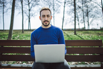Fototapeta premium Young man in blue sweater in a park teleworks with a computer in a park and looks at the camera