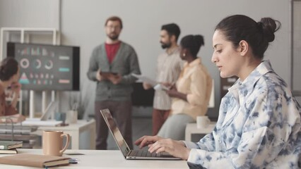 Side view waist up slowmo of young Middle Eastern woman typing on laptop at office desk while her diverse colleagues making product presentation in blurred background