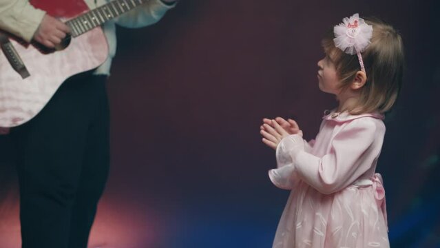 Little girl on stage in pink vintage dress, she claps his hands and dances, her father plays acoustic guitar. Color music is shining. Performance on stage. Early development of children