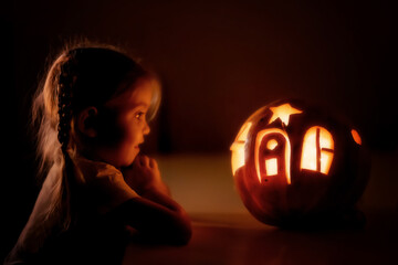 Happy excited little girl looking inside of glowing jack o lantern, carved pumpkin, while standing...