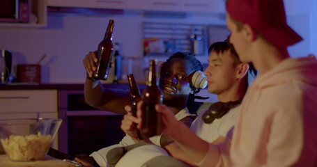 Celebrate victory while watching TV. Three multi ethnic guys clink bottles of drink, congratulate each other. Chips on the table in bokeh. Blue background of the evening flat.