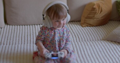 Cute little girl sits with headphones on her head, listens to sounds, smiles. Child exposed to...