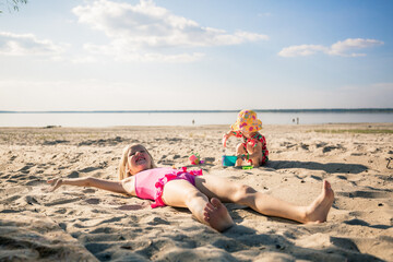 A 7 year old girl is lying on the beach and sunbathing in a pink bathing suit. Little 3 year sister...