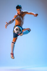 Portrait of young male football player in motion, dribbling, kicking ball isolated over blue background in neon light