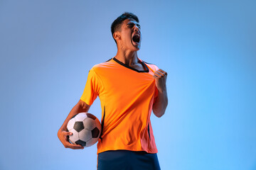 Portrait of young expressive man, football player posing isolated over blue studio background in...