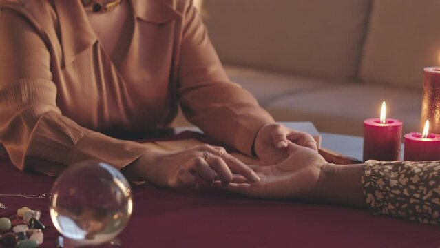 Cropped slowmo of unrecognizable female fortune teller conducting palm reading of woman sitting in front of her at table in dark room with burning wax candles