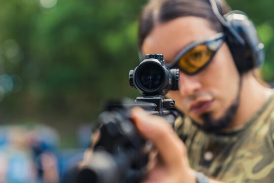 White man in safety goggles and headphones wearing camo t-shirt aiming submachine gun on firing range. Gun sight. Horizontal shot . High quality photo