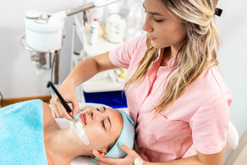 Beautician therapist applying face treatment in beauty salon to young woman.