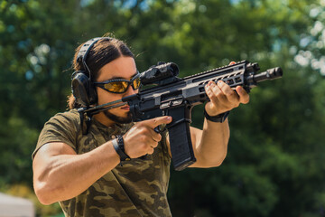 Fototapeta premium White muscular man in camouflage t-shirt and safety gear firing submachine gun on training ground. Outdoor horizontal shot. High quality photo