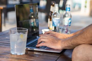 Side view, male hands type on a laptop on a wooden table, next to a glass with a refreshing drink, in a relaxed atmosphere.