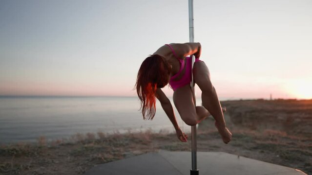 Athletic Woman in a Pink Tracksuit Performs Tricks on a Pole on Background Sea. Female Pole Dancer Does Acrobatics. Sports and Healthy Lifestyle. Slow Motion.