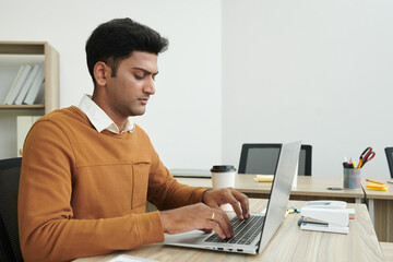 Serious young Indian businessman working on laptop at office desk