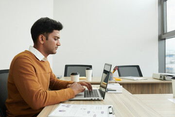 Concentrated young Indian entrepreneur working on laptop in modern office, checking reports and e-mails