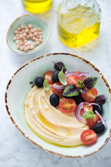 Green bowl with hummus, cherry tomatoes, black olives, red onion and basil leaves, middle closeup on a light-grey marble background